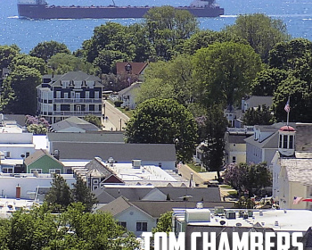 A ship sails on blue waters in the background, while residential buildings and lush green trees occupy the foreground. Text overlays the image, reading "Memories of a Mackinac Island Native" and "Tom Chambers.