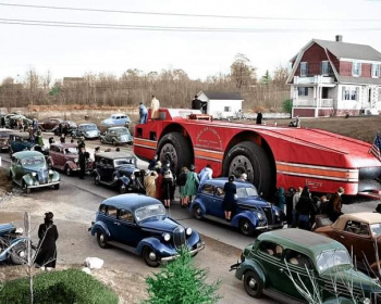A large futuristic-looking vehicle with multiple wheels is parked on a road, attracting a crowd of people. Vintage cars surround it, and spectators gather to look. A house and trees are in the background.