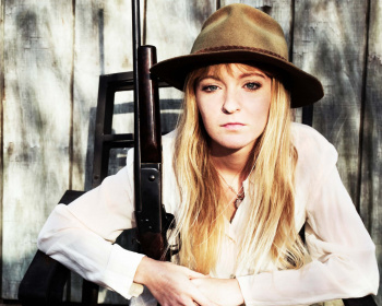 A woman wearing a hat and torn jeans sits on a chair holding a rifle in front of a wooden fence. Above her, the title "Roadkill Justice" is in bold, distressed red letters. The name "Terri Martin" is written at the bottom in white text.