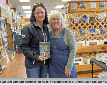 Two women stand side by side in a bookstore. The woman on the left holds a book titled "Rescuing Crash." Books and wooden shelves are visible in the background. Both women are smiling at the camera.