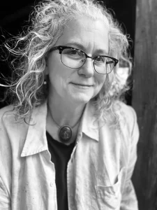 A person with curly, gray hair and glasses wears a light-colored button-up shirt over a dark top and a large pendant necklace. They are looking at the camera and smiling slightly. The photo is in black and white.