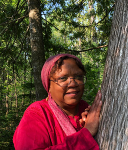 A woman wearing glasses, a red headscarf, and a red shirt stands outdoors in a forest, resting her hands against the trunk of a large tree. Sunlight filters through the trees, illuminating her face.
