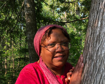 A woman wearing glasses, a red headscarf, and a red shirt stands outdoors in a forest, resting her hands against the trunk of a large tree. Sunlight filters through the trees, illuminating her face.