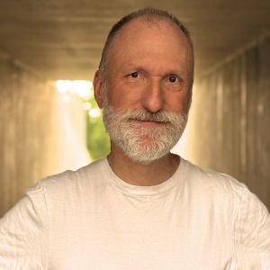A middle-aged man with a gray beard and short hair stands in a tunnel, wearing a plain white t-shirt. Sunlight shines from the background, softly illuminating his face.